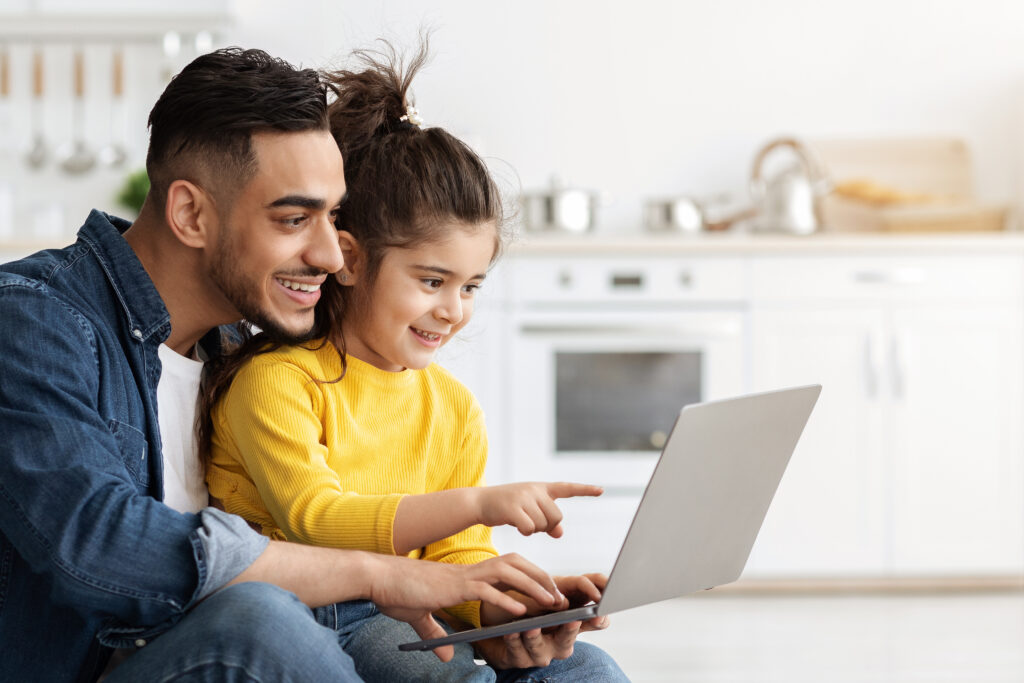 Online Shopping Concept. Happy Arab Dad And Little Daughter Using Laptop Computer Together While Relaxing In Kitchen At Home, Cute Little Girl And Middle Eastern Father Enjoying Internet Purchases